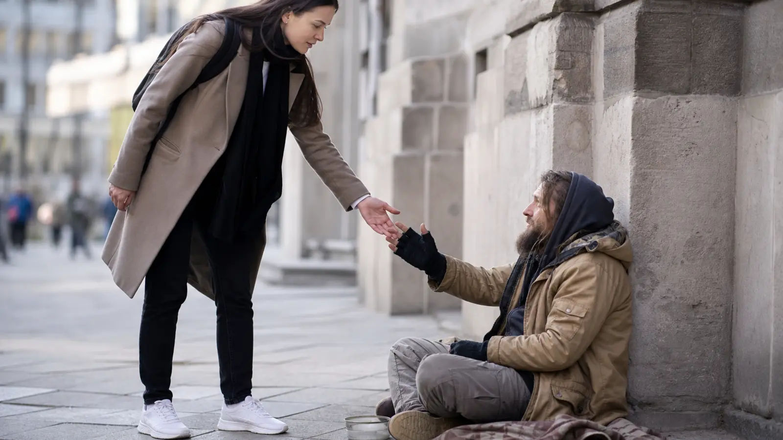 Eine Frau reicht einem sitzenden Mann auf einem Stadtgehweg die Hand.
